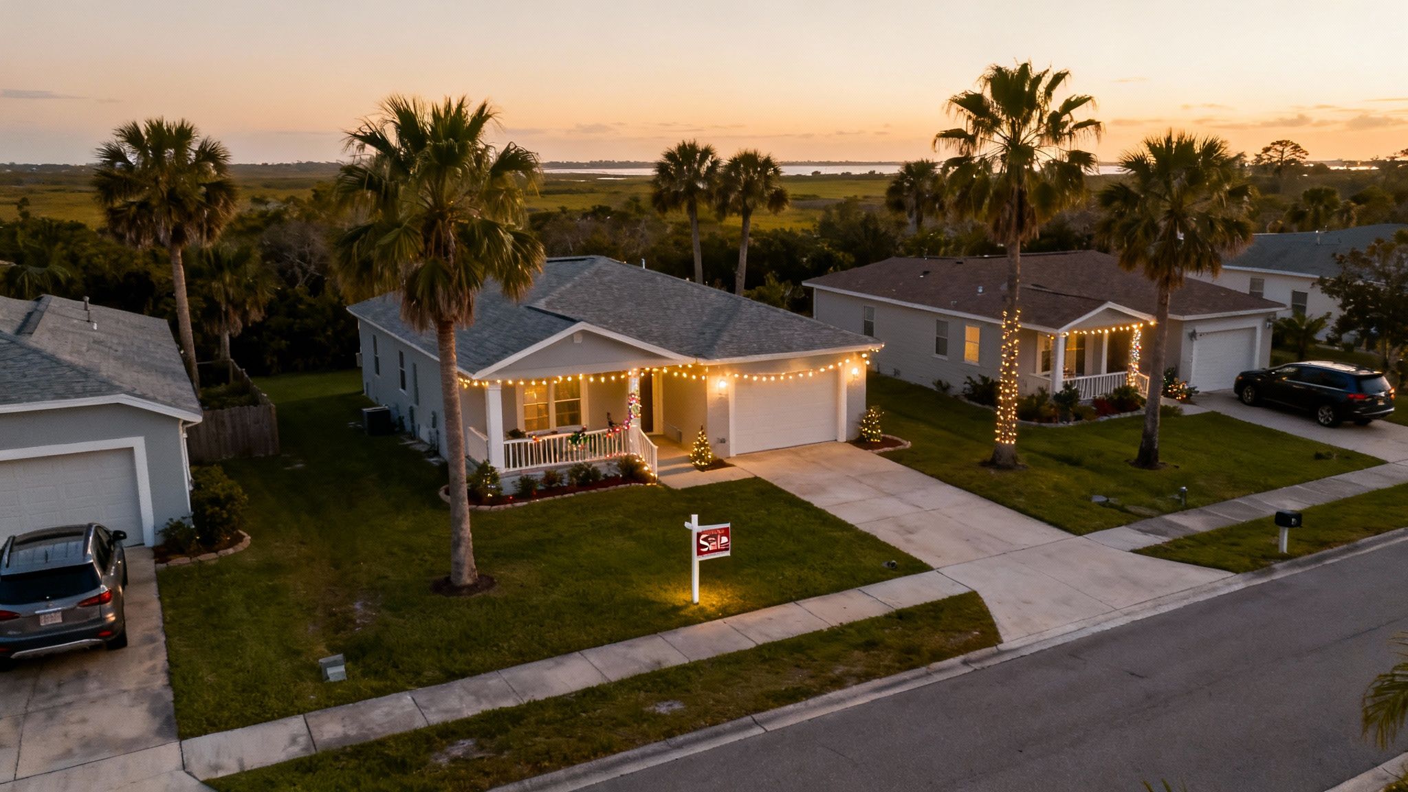 An aerial view of suburban houses decorated with holiday lights, one with a for sale sign, at sunset.