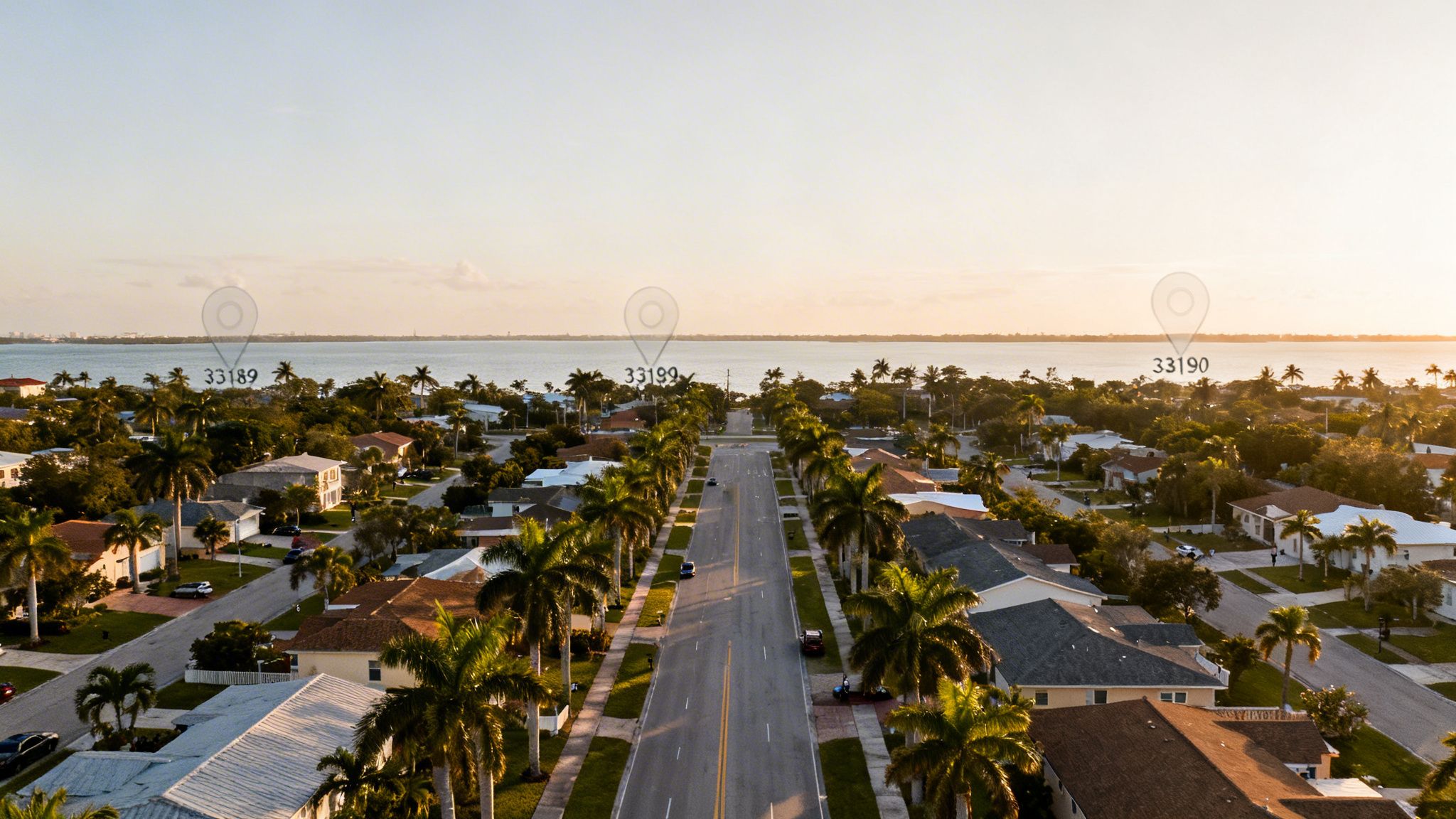 Aerial view of a palm-lined residential street leading to a bay at sunset, showing houses and zip codes.