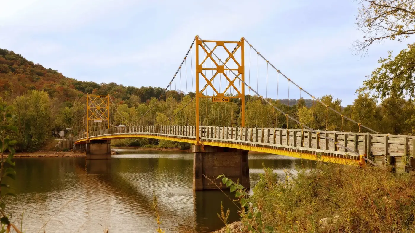 A narrow yellow suspension bridge with a timber deck crossing a still river, with wooded hills in autumn colour rising behind it