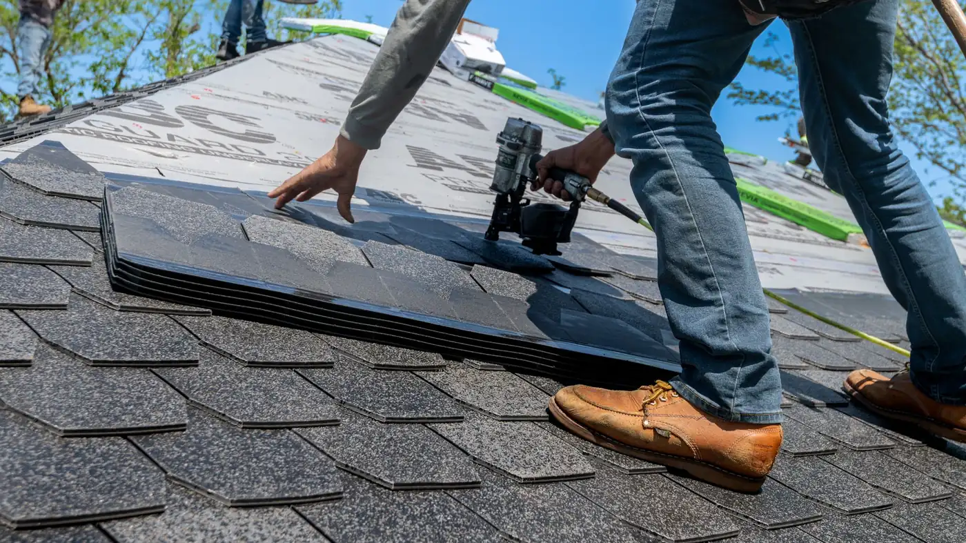 A roofer in work boots kneeling on a half-finished roof, nail gun in hand, laying a course of grey asphalt shingles over the underlay