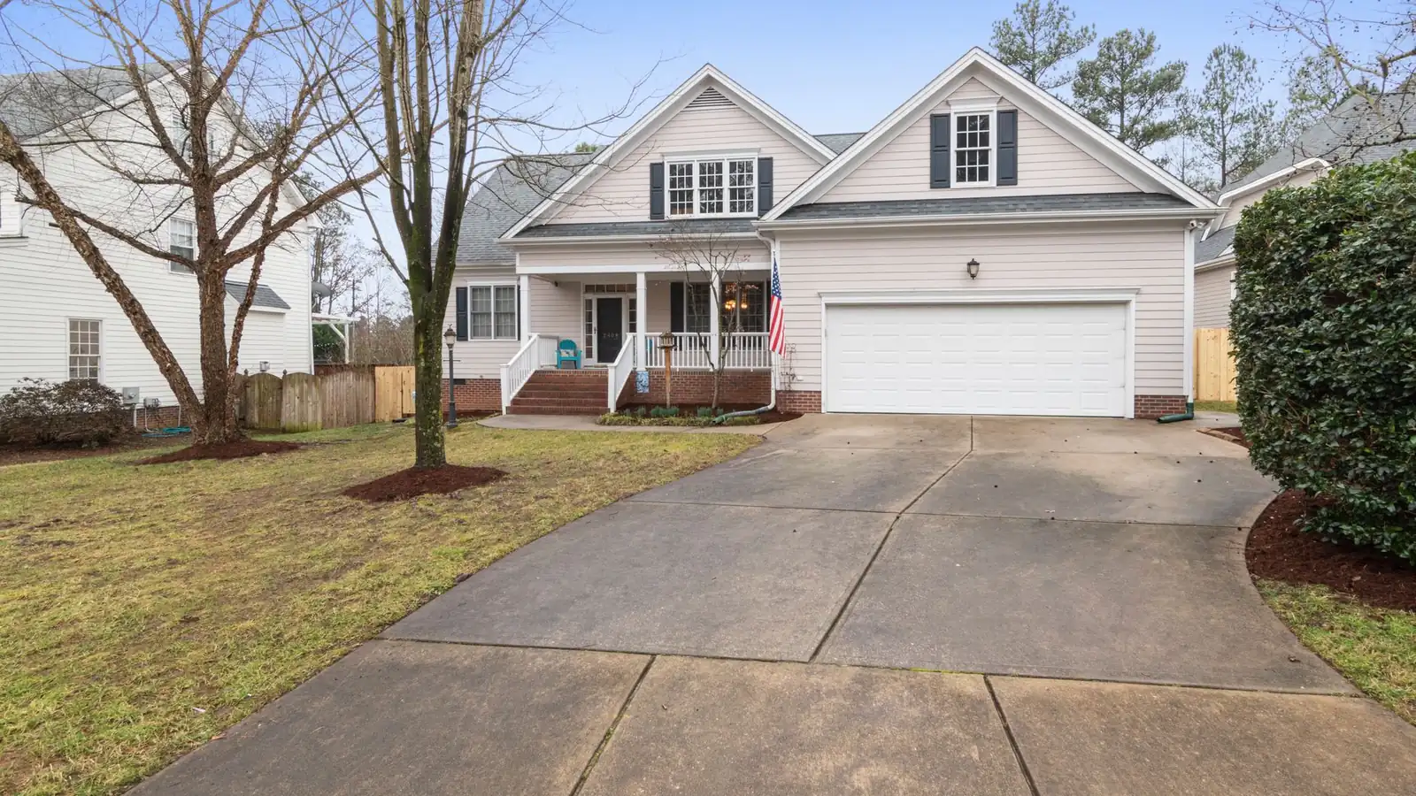 A cream two-storey house with dormer windows, a covered front porch and a flag by the door, at the top of a wide concrete driveway with bare winter trees around it