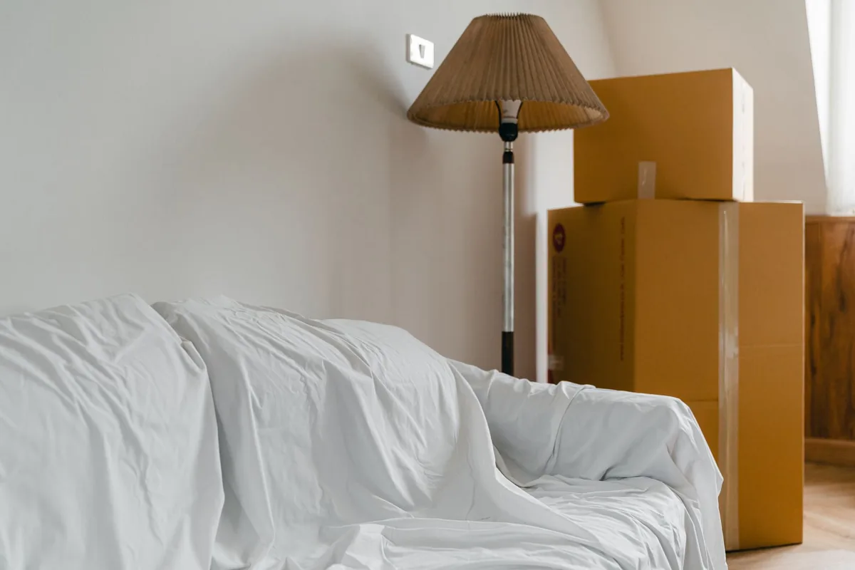 Sofa covered with a white sheet next to stacked cardboard boxes and a floor lamp