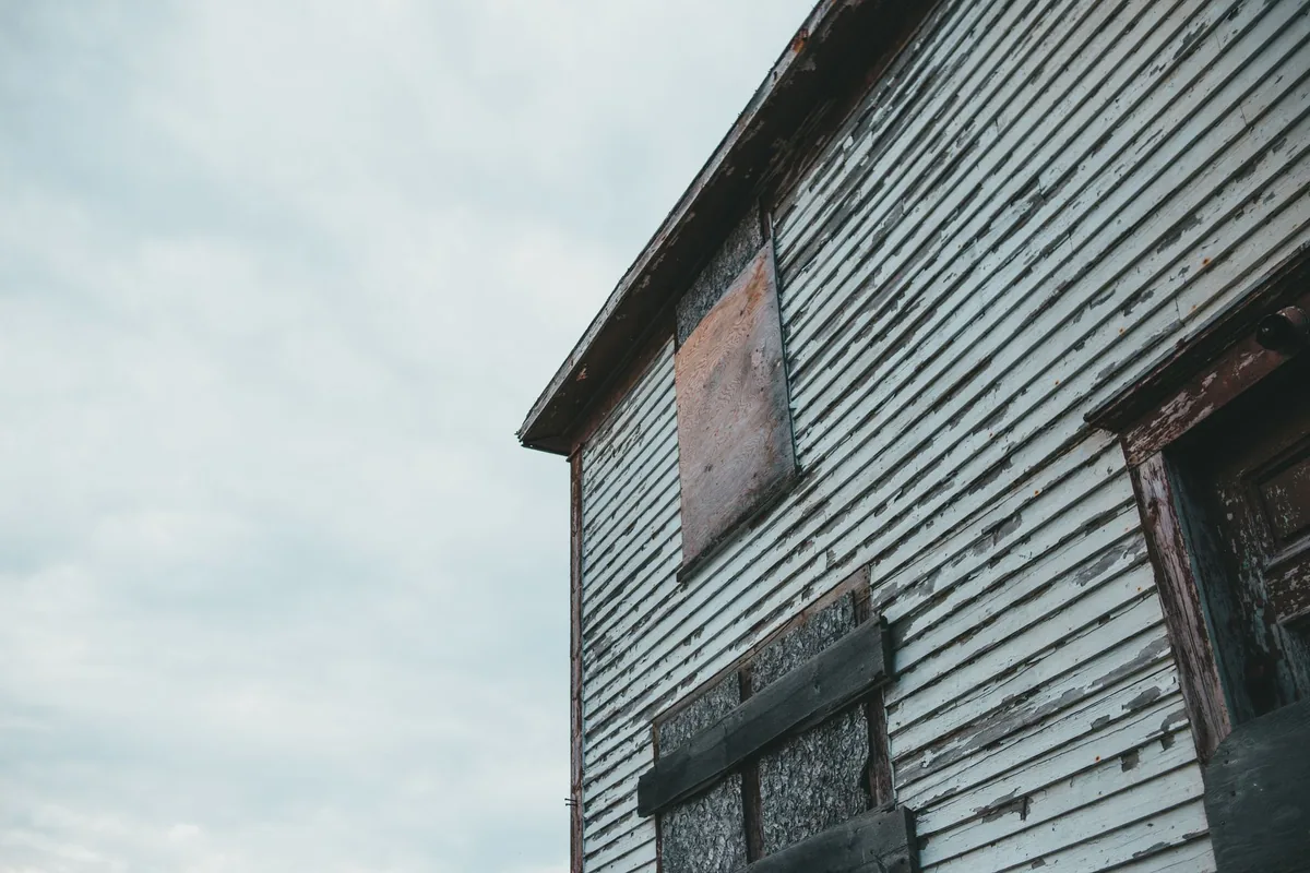 Boarded-up wooden house with peeling white paint under a cloudy sky
