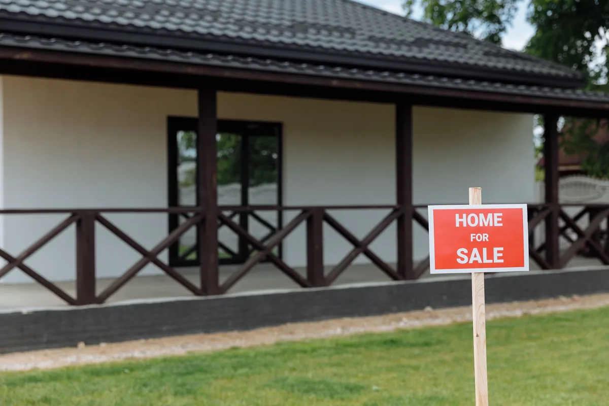 Red Home for Sale sign on a lawn in front of a white house with a covered porch