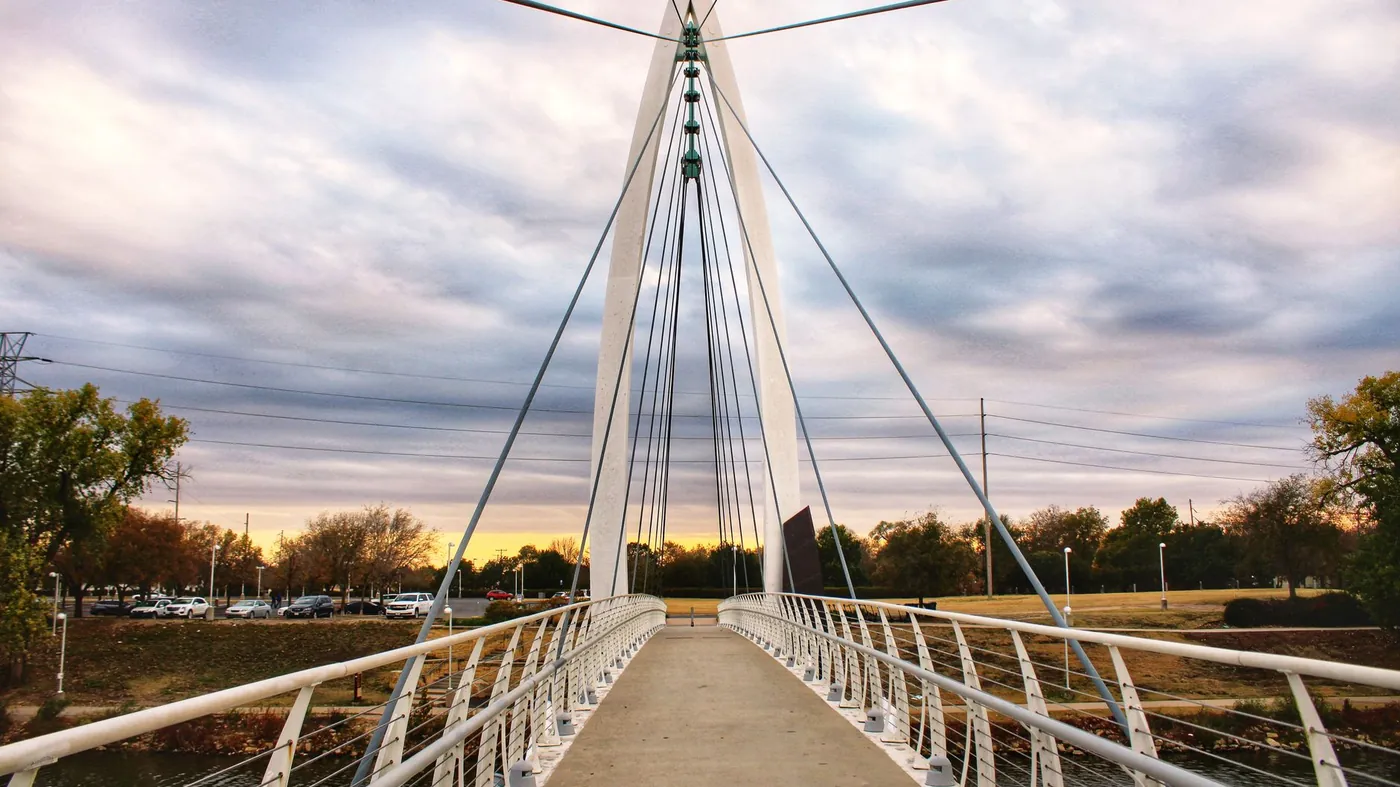 A white cable-stayed pedestrian bridge with a tall pointed pylon crossing a river at sunset, with parkland and a low tree line beyond