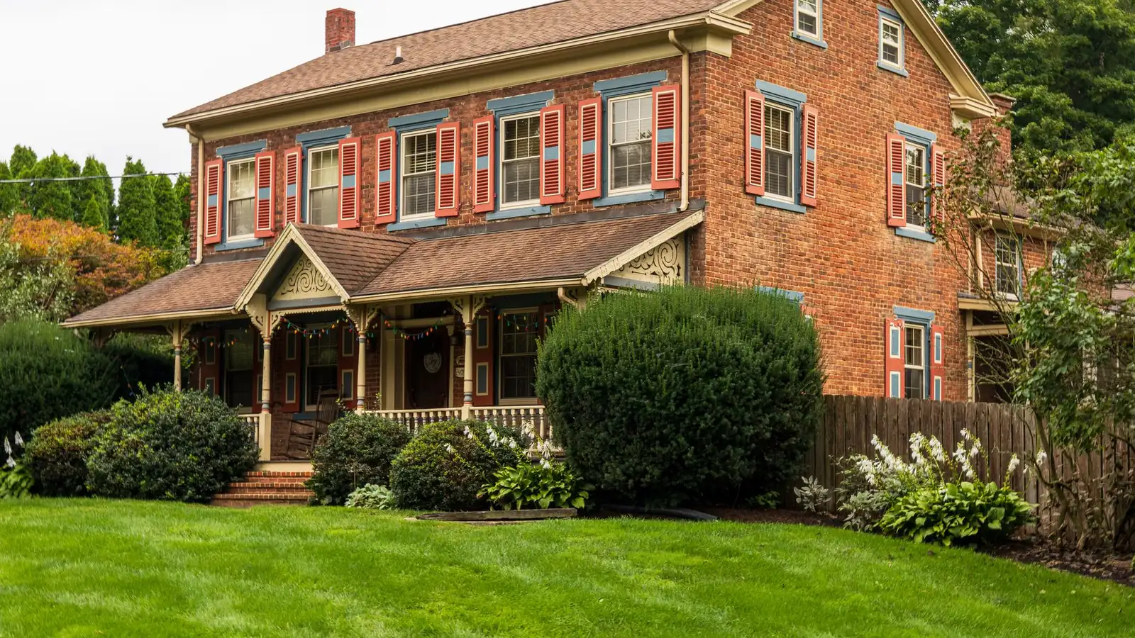 An older two-storey red brick house with painted shutters, a decorated porch with turned posts and clipped box hedges across the front lawn