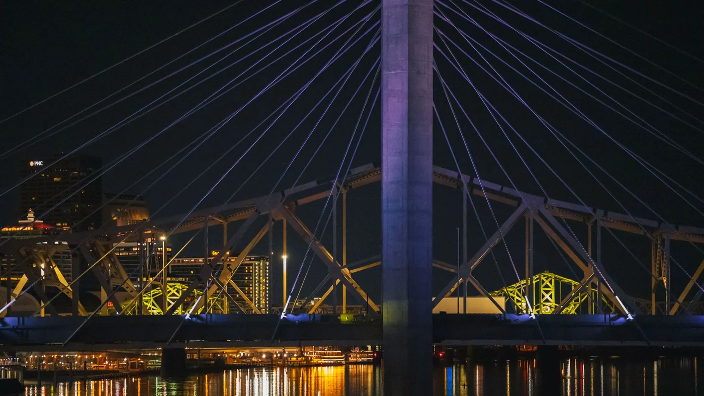 The Louisville riverfront at night, with the lit cables of the downtown bridge, the yellow steel of the older truss spans behind it and the city reflected in the Ohio River