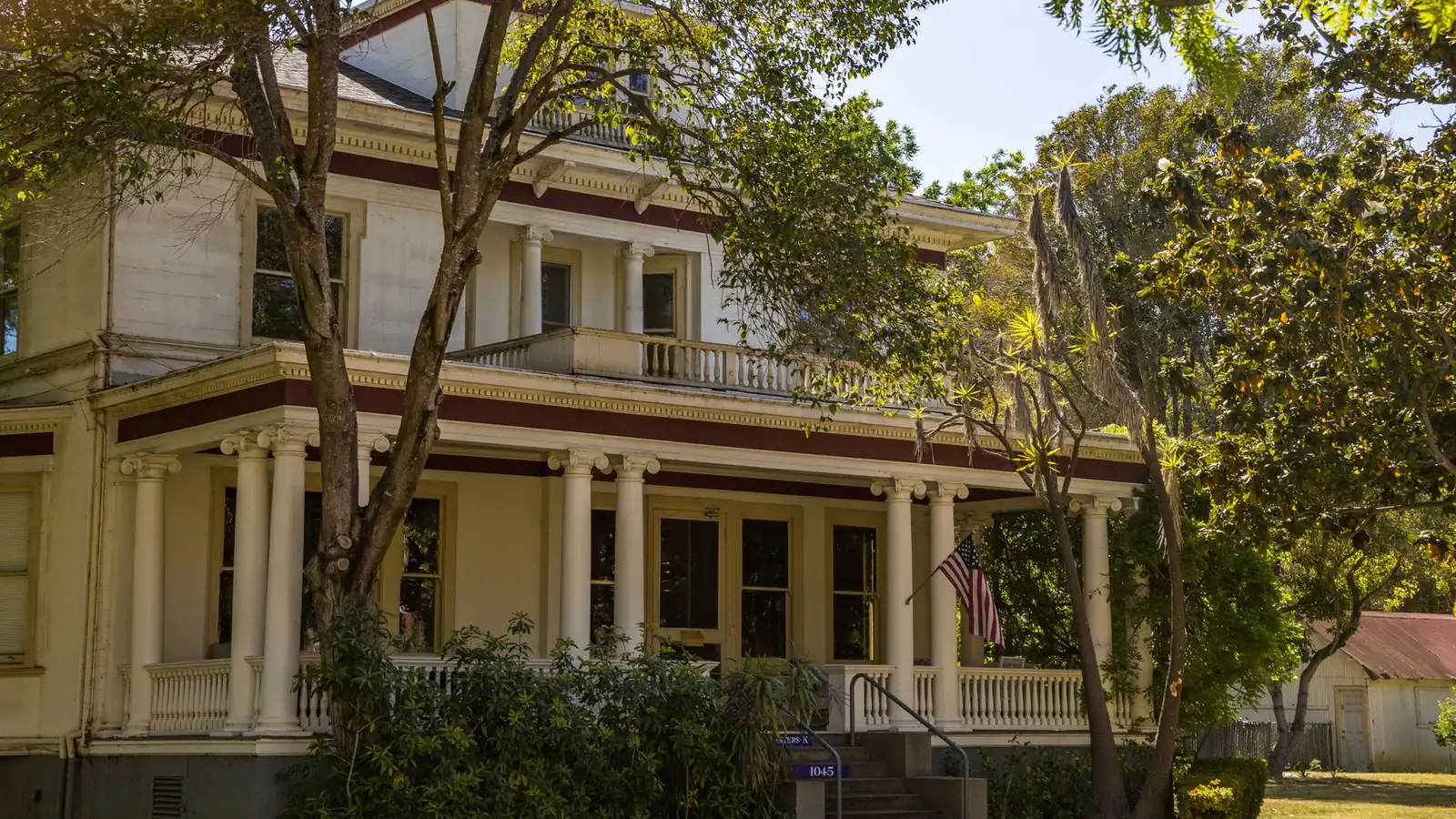 A large older frame house painted cream with deep two-storey porches, turned columns and a balustraded upper gallery, half shaded by mature trees