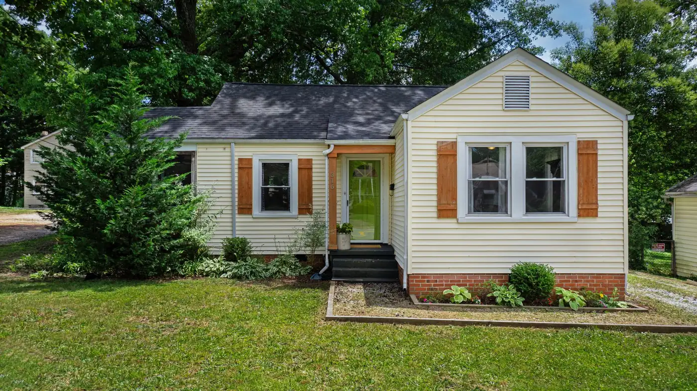 A small cream weatherboard cottage with cedar shutters, a brick skirt, front steps and a planted bed, on a shaded lawn