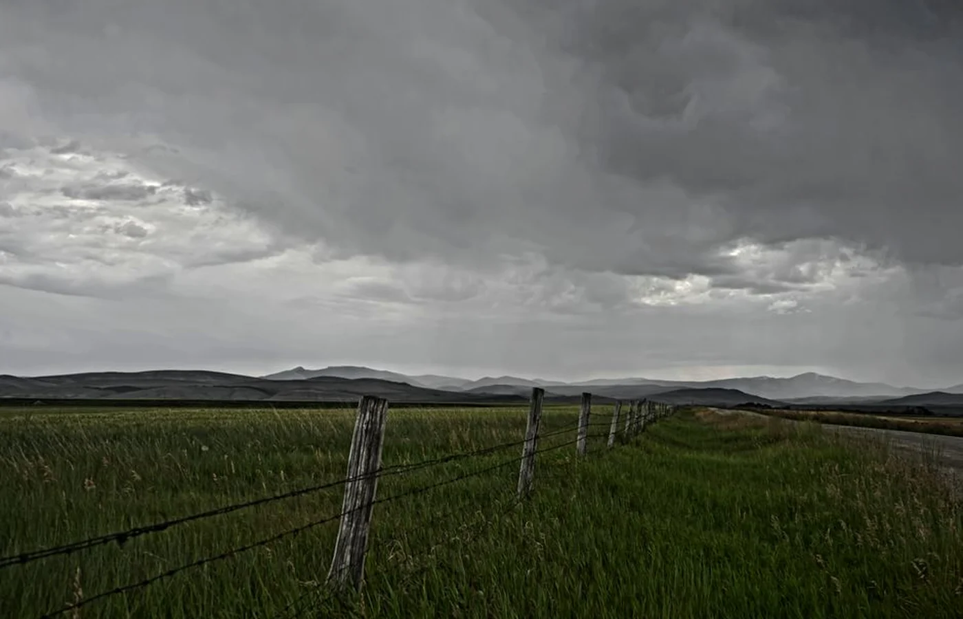 A rural Montana landscape under heavy overcast, with a barbed wire fence running along dry rangeland and mountains dark on the horizon