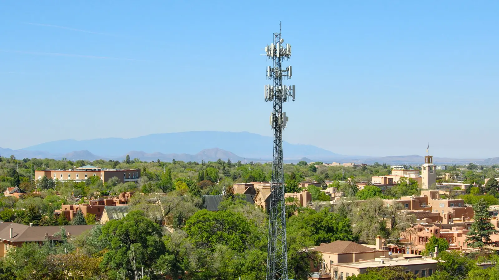 Santa Fe seen from above, with flat-roofed adobe buildings in soft earth colours among cottonwoods, and low blue mountains along the horizon