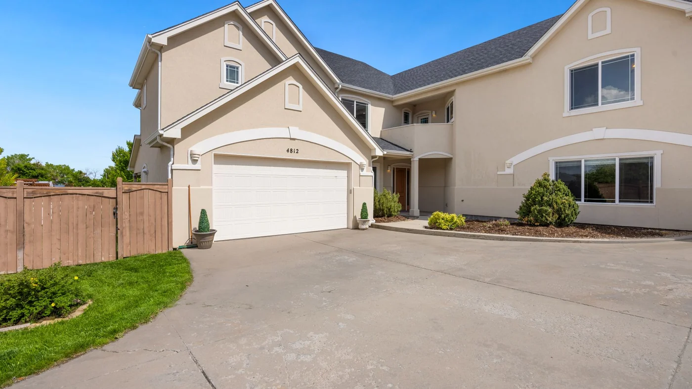 A tan stucco two-storey house with arched detailing over a white double garage door, a broad concrete drive and clipped shrubs in gravel beds