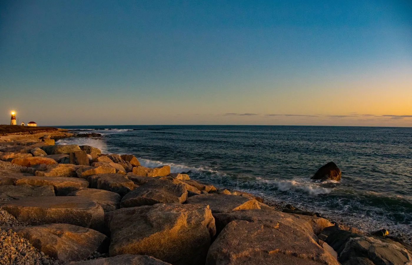 A rocky Rhode Island shoreline at sunset with a lighthouse on the point and low light across the water