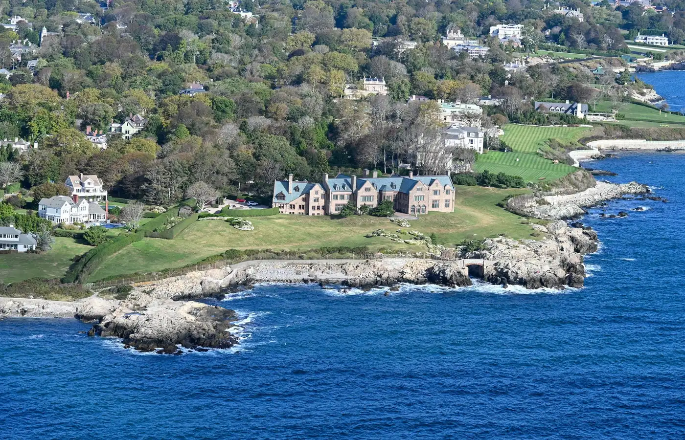 An aerial view of the Newport coastline, with large houses and lawns on a green headland above a rocky shore and deep blue water
