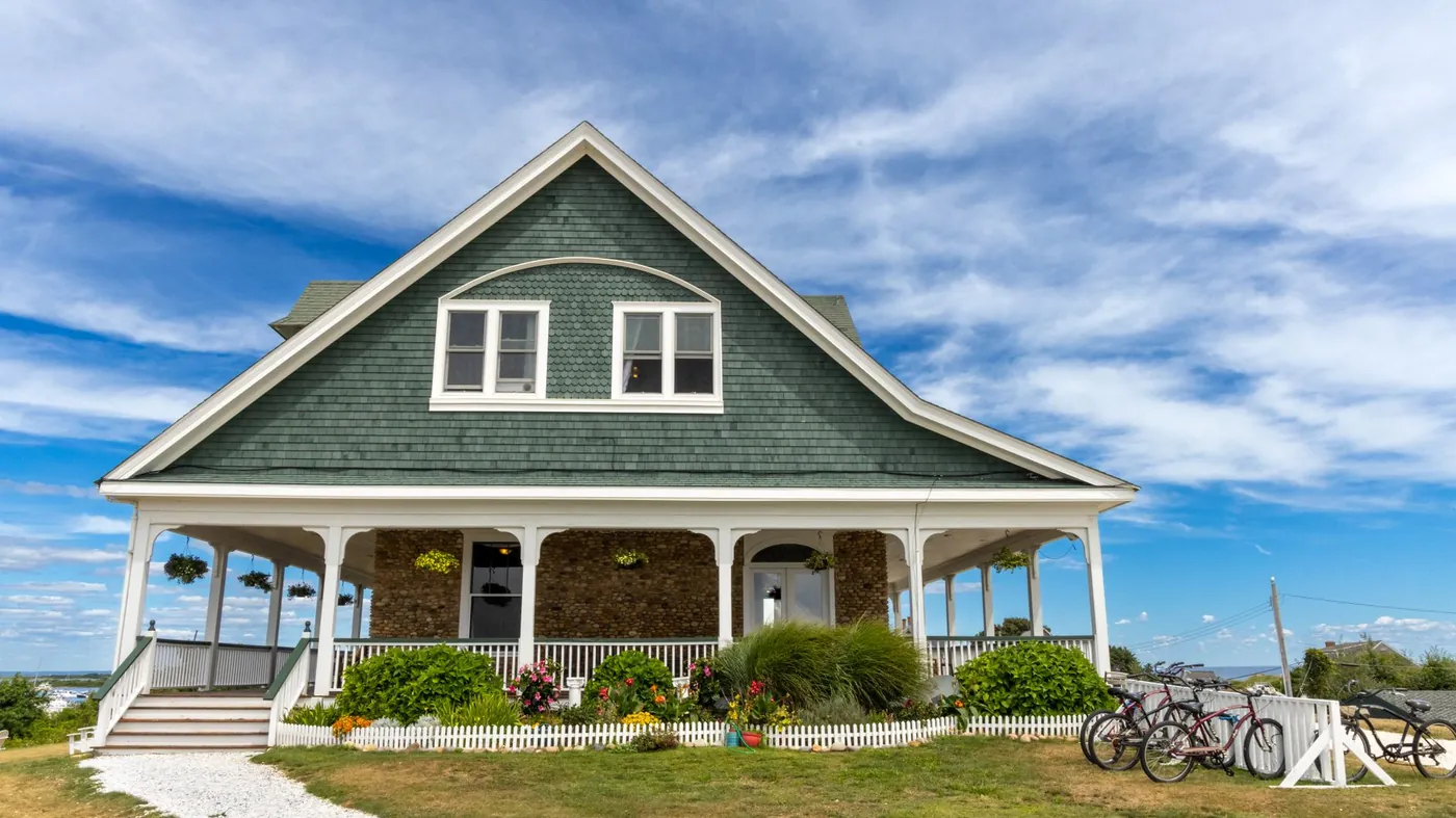 A shingled house behind a low hedge and a flower garden on a bright day, with bicycles leaning against the fence and a blue sky above