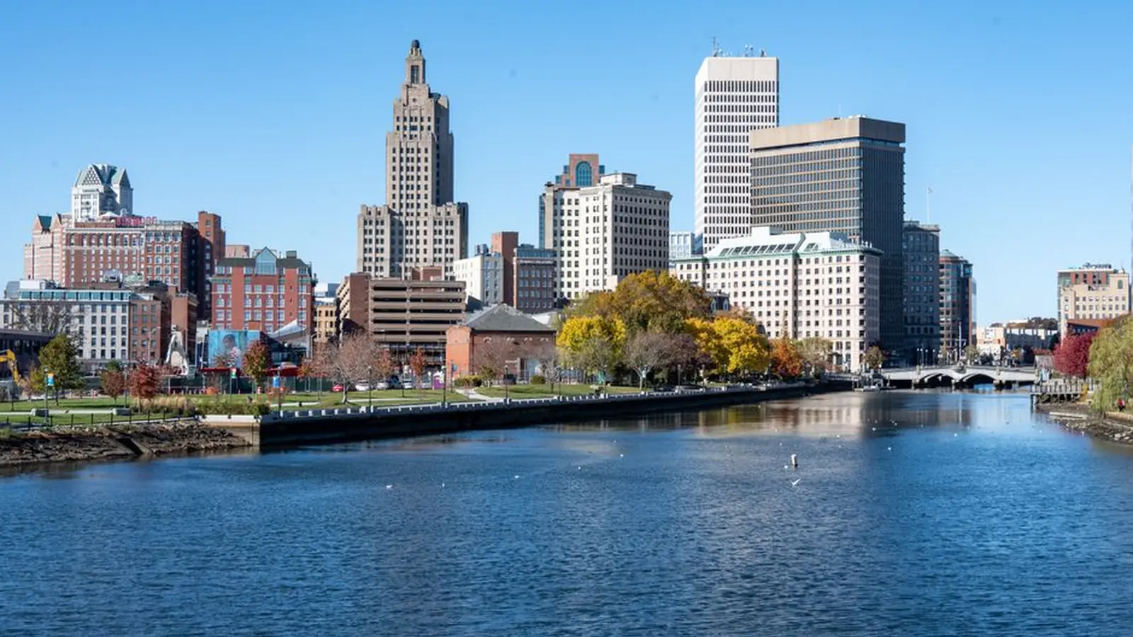 Downtown Providence seen across the river on a clear autumn day, with the Industrial Trust tower and a cluster of mid-century office blocks above a tree-lined riverwalk