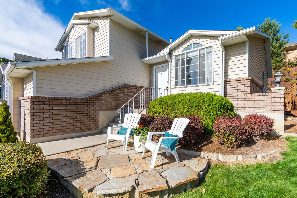 Tan suburban house with a brick wall, stone patio and two white chairs out front