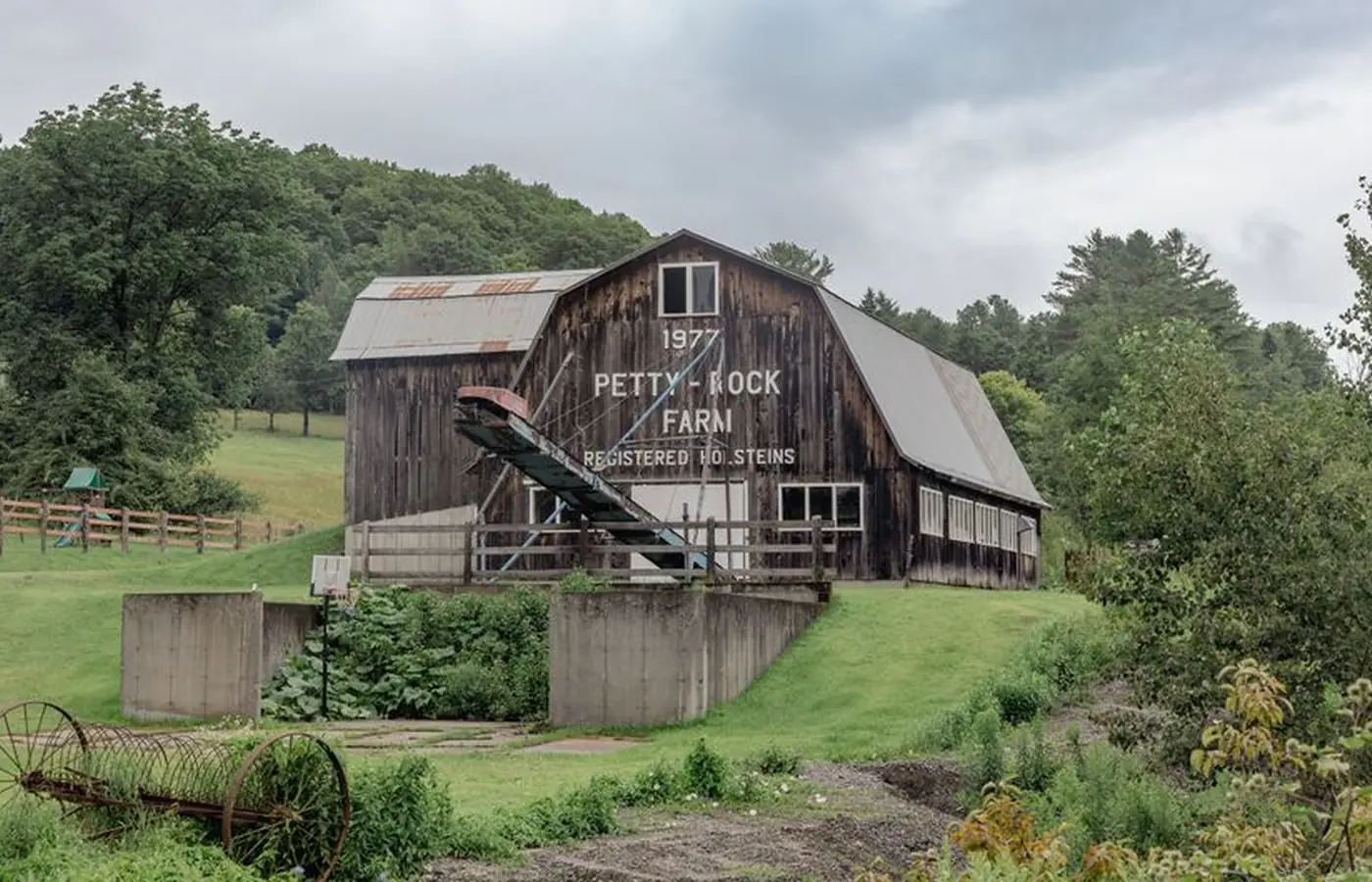 A weathered wooden barn standing in green rolling countryside in Vermont, with trees and hills running back behind it