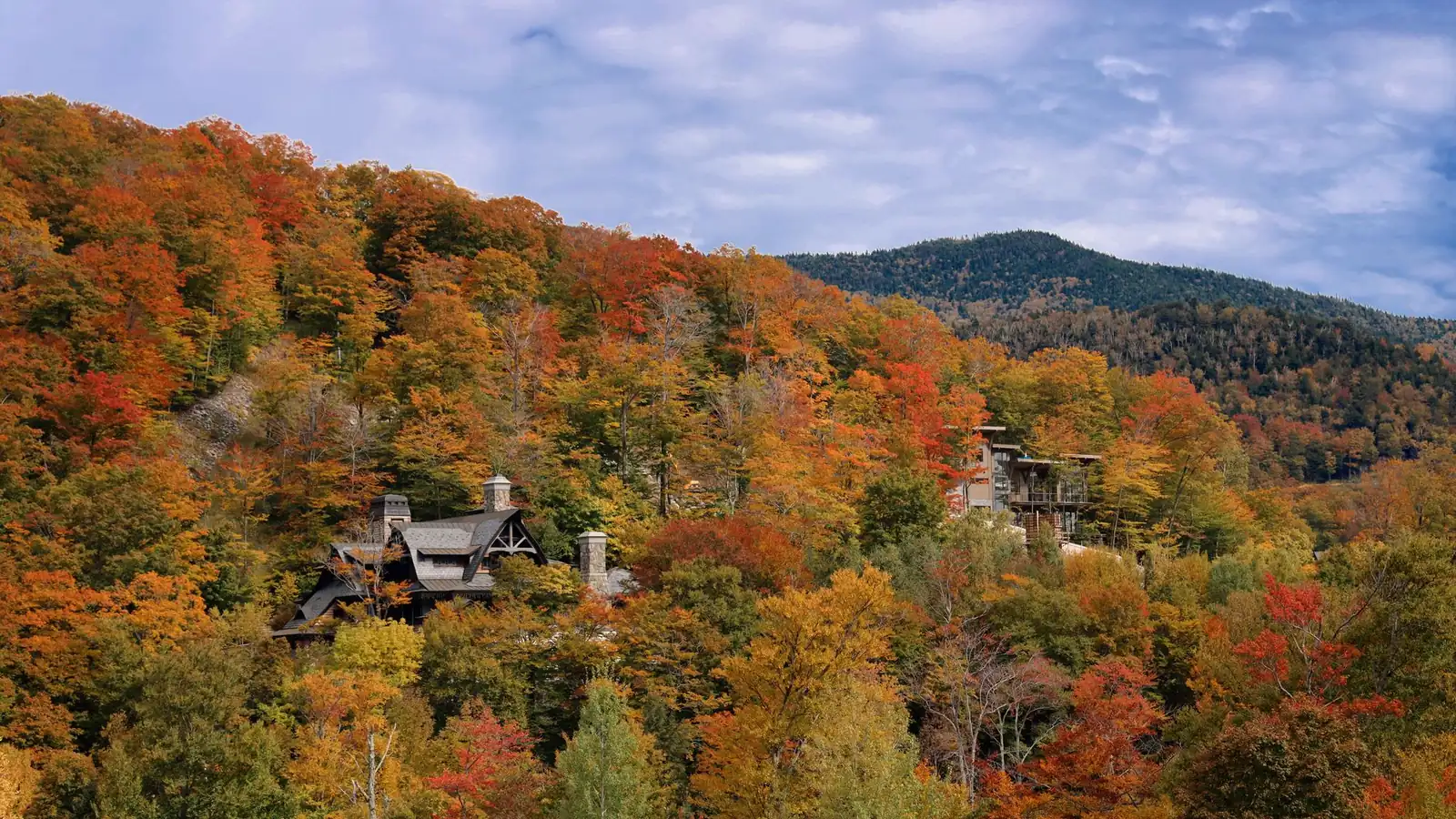 Houses set into a steep hillside at Stowe among maples in full autumn colour, with a forested green ridge rising behind them under a broad cloudy sky